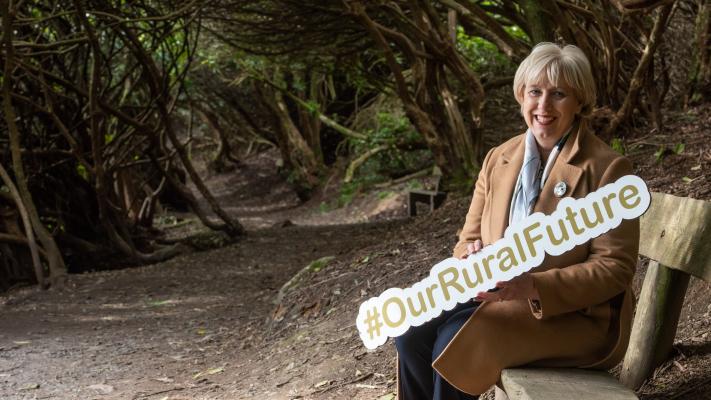 Minister Heather Humphreys holding a #OurRuralFuture sign. She is seated in front of a woodland background.