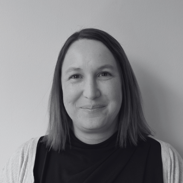 Black and white portrait of Ann Marie Spillane with shoulder-length hair, wearing a dark top and light cardigan, standing against a plain background.
