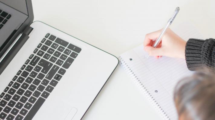 An image of a woman writing notes while looking at a computer screen.