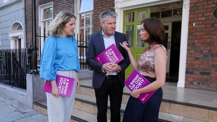 Three individuals holding purple covered paper documents standing outside the red-bricked façade of Buswells Hotel in Dublin city centre. Fiona Coyle of Mental Health Reform, Ivan Cooper of The Wheel, and Pauline McKeown of Coolmine Therapeutic Community outside The Wheel's Pre-Budget Submission Briefing for Budget 2024.