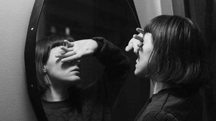 Black and white image of the head and shoulders of a woman with medium length cropped hair close up to and facing a large oval mirror with her hand held over her eyes palm out. 