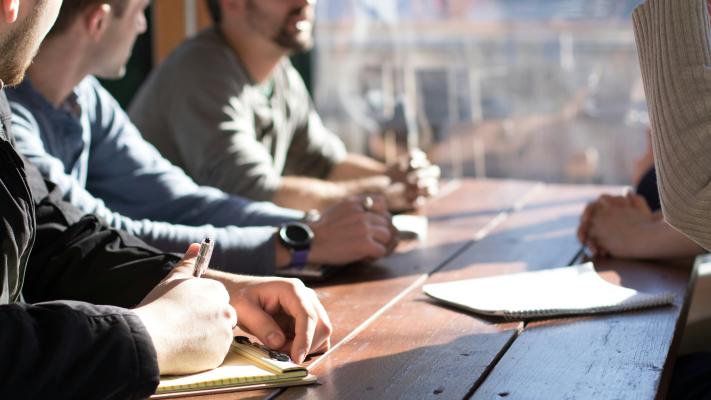 Several male figures in business casual dress seated around a wooden table taking notes and engagine in discussion. 