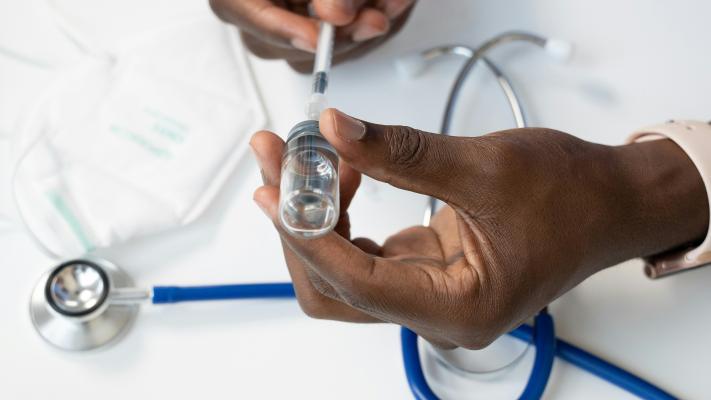 A close-up image of a pair of hands drawing medicine from an ampoule with a hypodermic needle above a blue stethoscope and facemask resting on a white table top.