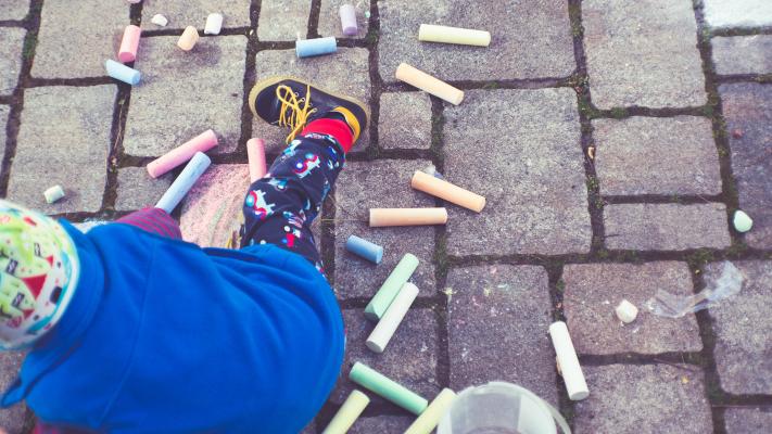 A small child in a blue pullover facing away from and below the camera sitting on a grey cobbled pavement littered with brightly coloured chalk.
