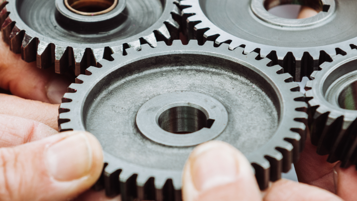 A close up image of three pairs of hands holding four dark grey steel cogs together. 