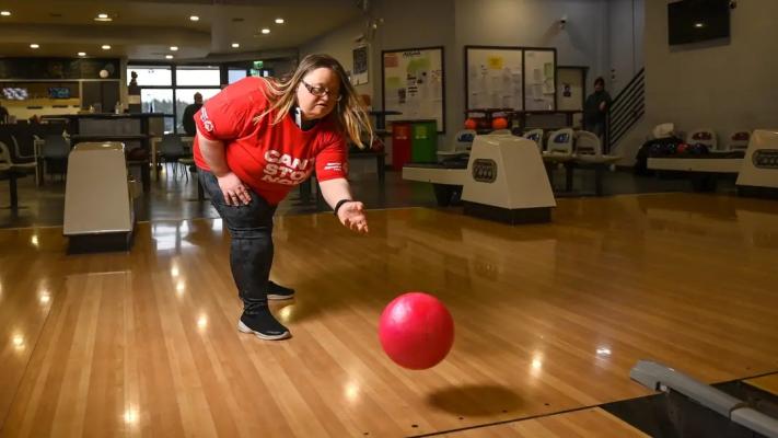 Image of a woman with blond hair in a red t-shirt ten-pin bowling with a red bowling ball. 