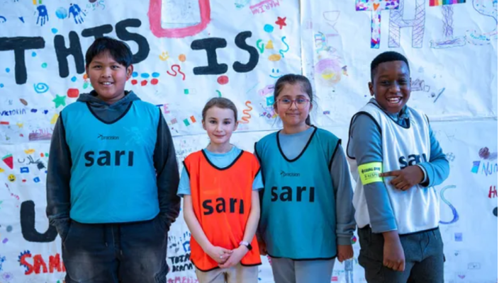 Four smiling children of diverse backgrounds stand side by side wearing colourful sports bibs with the word “sari” printed on them. Behind them is a large, hand-decorated banner reading “THIS IS US” surrounded by drawings and doodles made by children.