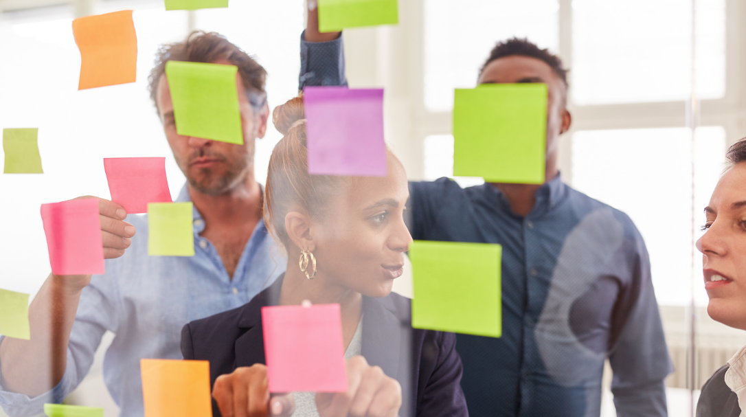 An image of two men and two women in casual business clothes working together with a wall of suspended coloured post-it notes.