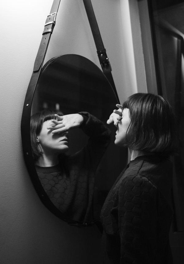 Black and white photo of the head and shoulders of a woman with mid length cropped dark hair close to and facing a large oval mirror with her right hand held across her eyes, palm out. 