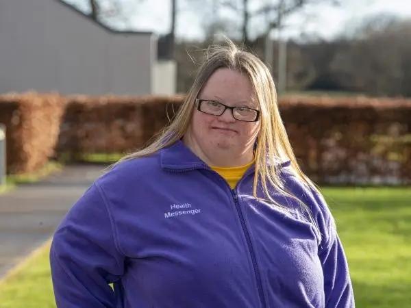 A woman with blond hair and glasses in a violet fleece top looking to camera standing in front of a lawn and hedges.