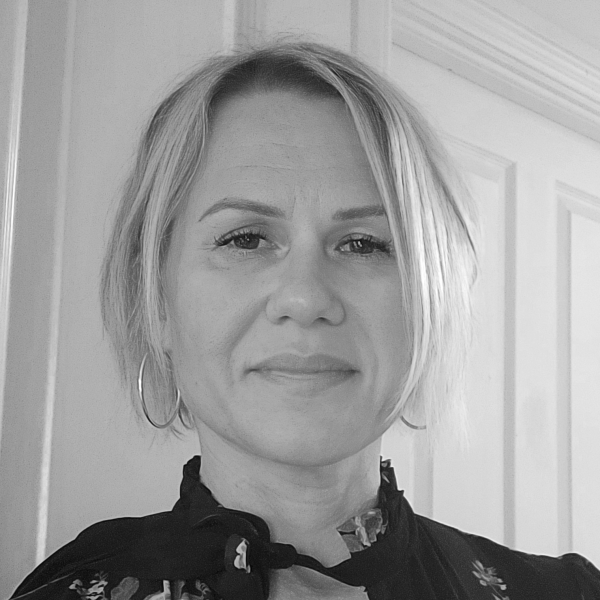 A person with light hair in a bob cut, wearing hoop earrings and a dark top with a high collar, stands in front of a white paneled door. The image is in black and white.
