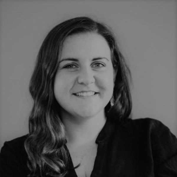 A woman with long wavy hair smiles at the camera. She is wearing a dark top and is posed against a plain background. The image is in black and white.