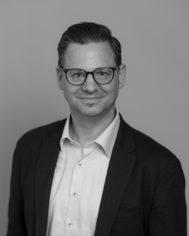 A man wearing glasses, a button-up shirt, and a blazer stands against a plain background. He has short, neatly-combed hair and a slight smile. The image is in black and white.