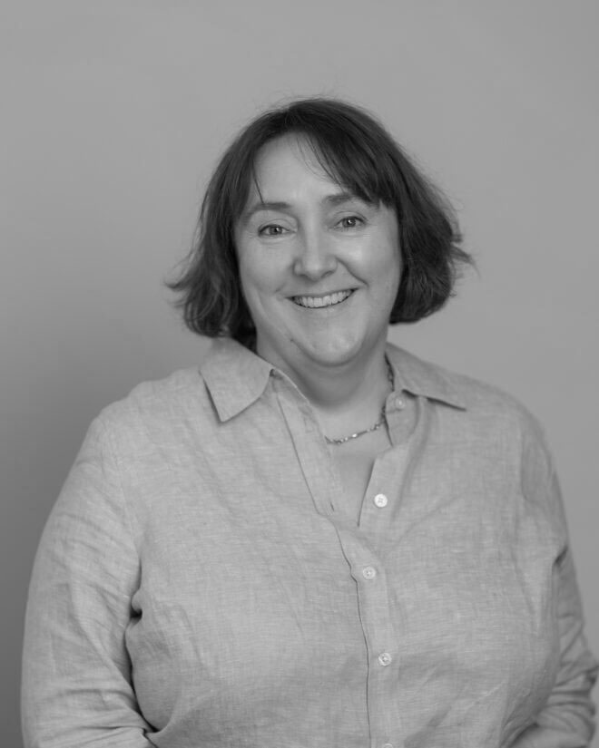 A woman with short hair wearing a button-up shirt and a necklace stands smiling at the camera against a plain background in a black and white photo.