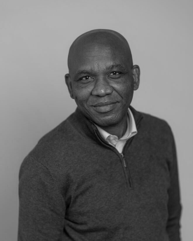 A man with a shaved head wearing a zip-up sweater and collared shirt stands against a plain background, looking at the camera and smiling slightly. The image is in black and white.