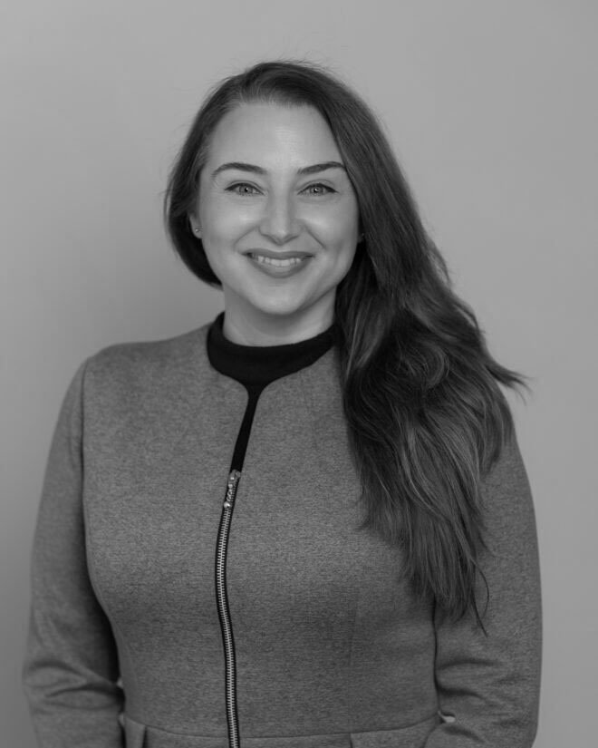 A woman with long hair wearing a zip-up jacket over a dark top stands in front of a plain background, smiling at the camera. The image is in black and white.
