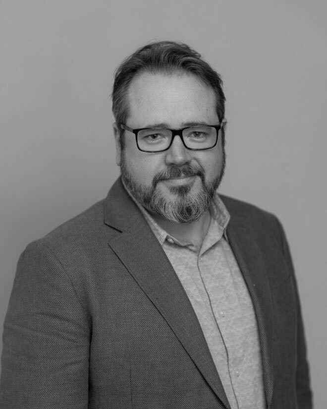 A man with glasses, a short beard, and mustache is wearing a light shirt and a blazer. He is facing the camera and smiling slightly. The image is in black and white with a plain background.