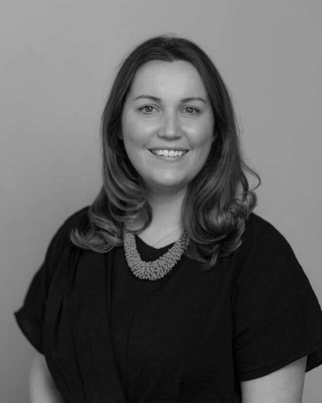 A woman with long hair wearing a dark top and a chunky necklace is smiling at the camera. The photo is in black and white, with a neutral background.