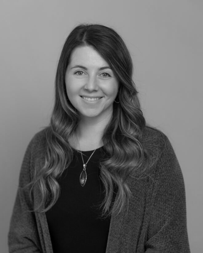 A woman with long wavy hair wearing a sweater, a dark shirt, and a pendant necklace smiles while facing the camera in a black-and-white studio portrait against a plain background.