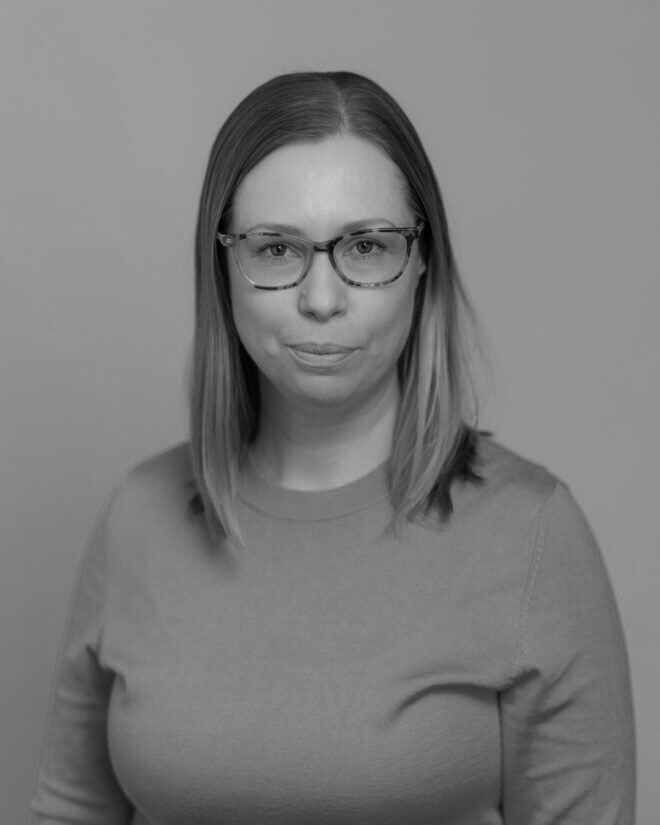 A woman with straight, shoulder-length hair and glasses faces the camera. She is wearing a plain, long-sleeve shirt. The image is in black and white with a neutral background.