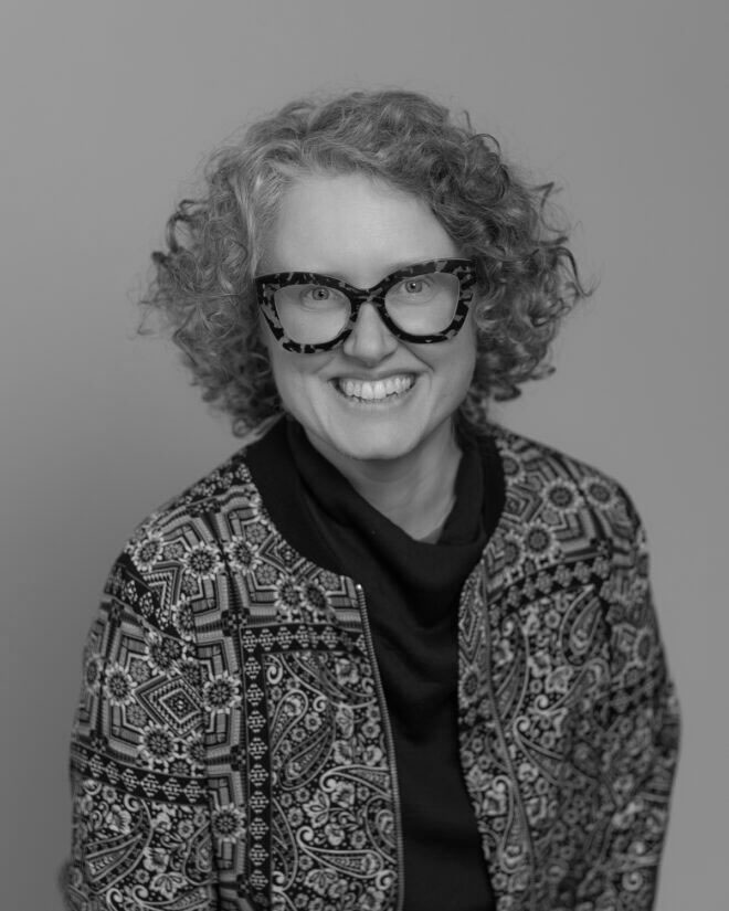 Black and white portrait of a smiling person with curly hair, wearing large patterned glasses and a patterned jacket over a dark top, posed against a plain background.