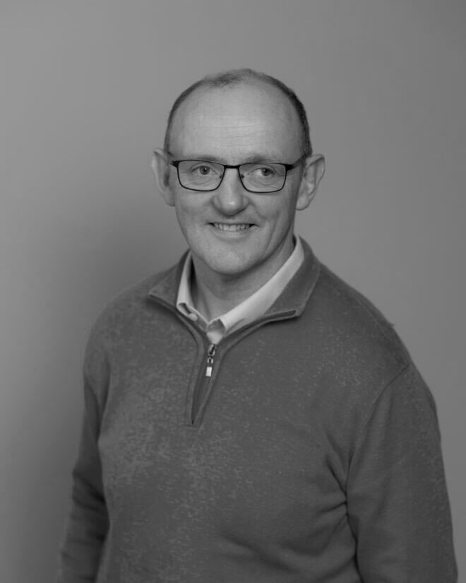 A middle-aged man with short hair and glasses is wearing a zip-up sweater over a collared shirt. He is smiling and looking slightly to the side against a plain background. The image is in black and white.