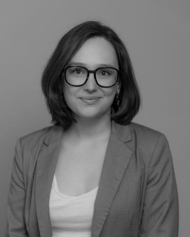 A person with shoulder-length hair and glasses wears a light-colored blazer over a white top, facing the camera and smiling slightly. The image is black and white with a plain background.