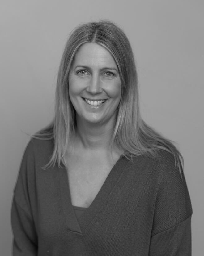 A woman with straight, shoulder-length hair smiles at the camera. She is wearing a V-neck top and is posed against a plain background. The image is in black and white.