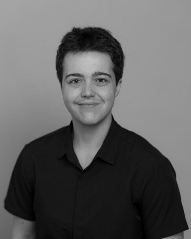 A person with short dark hair, wearing a dark collared shirt, is facing the camera and smiling gently. The background is plain and light-colored. The image is in black and white.