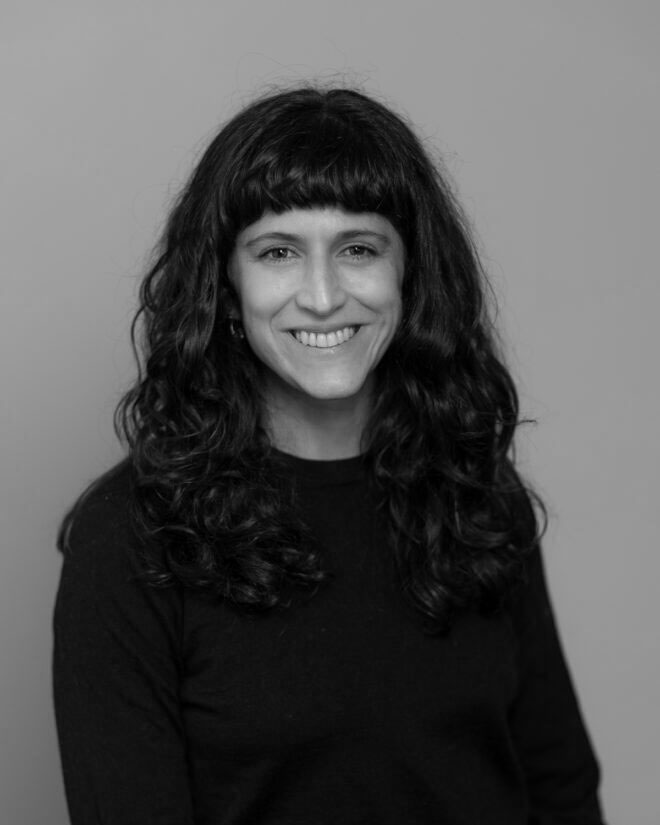 A woman with long, wavy dark hair and bangs is smiling at the camera. She is wearing a dark long-sleeve top and is posed in front of a plain, light background. The photo is in black and white.