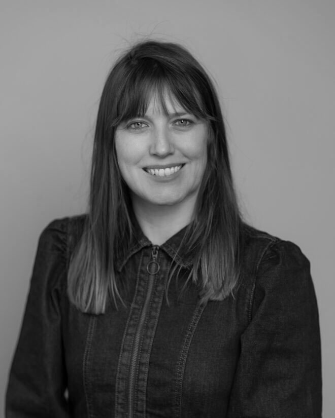 A woman with straight, shoulder-length hair and bangs is smiling at the camera. She is wearing a dark, long-sleeve, zip-up top. The background is plain and light in color. The photo is in black and white.