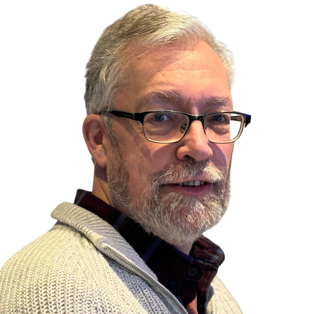 A man with gray hair, a beard, and glasses wears a light gray sweater over a dark shirt. He is facing slightly to the side and smiling, with a neutral white background.