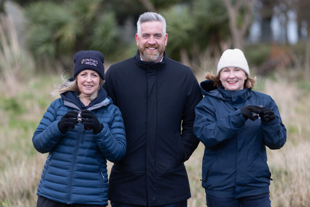 Three adults wearing winter jackets stand outdoors in front of greenery. Two women, one on each side, wear hats and gloves; a man stands in the middle. All three are smiling at the camera.