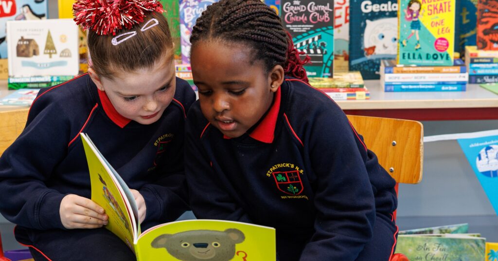 Two young girls in dark school uniforms sit closely together on a wooden chair, reading a childrens book. Colorful books are displayed on shelves behind them in a classroom or library setting.