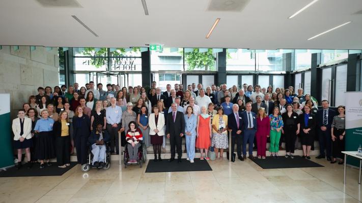 A large group of people in formal and semi-formal attire pose for a group photo in a modern, well-lit lobby with glass doors and windows in the background.