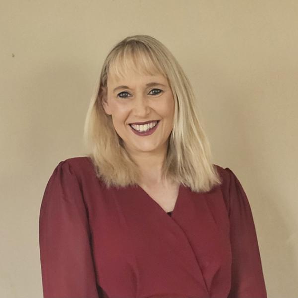 A woman with straight blonde hair and bangs is smiling at the camera. She is wearing a long-sleeved, dark red top and standing against a plain beige background.