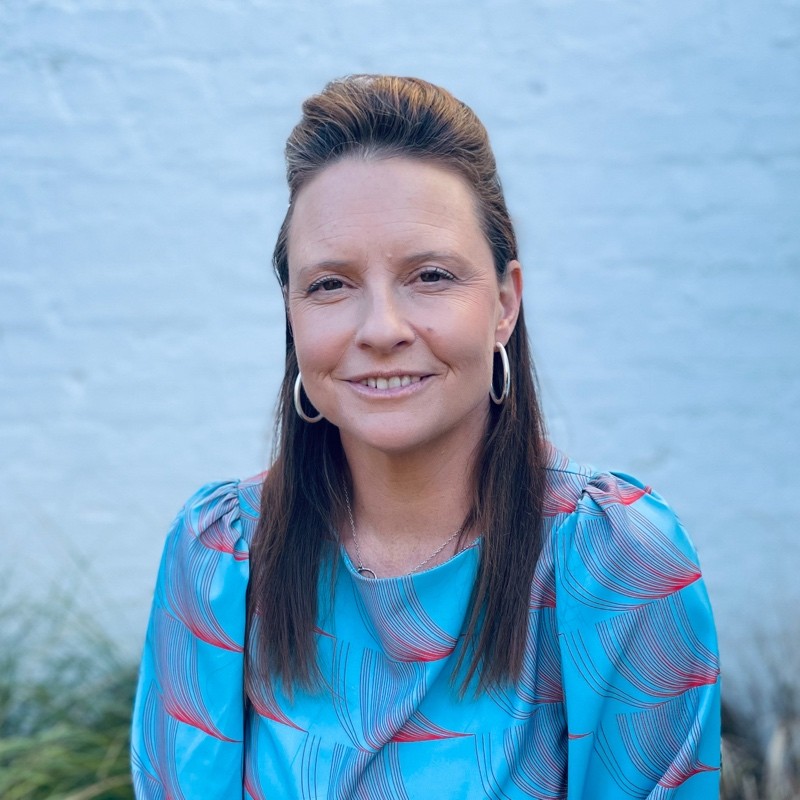 Woman with straight brown hair and hoop earrings wears a blue top with a red and white pattern. She is seated and smiling slightly, with a blurred light blue brick wall and some greenery in the background.