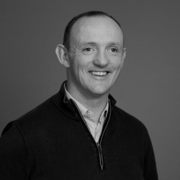 A man with short hair smiles at the camera. He is wearing a zippered sweater over a collared shirt. The background is plain and the photo is in black and white.