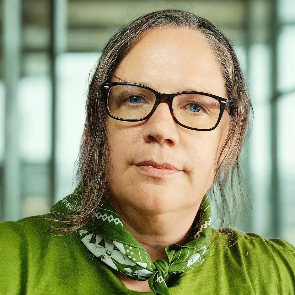 A person with straight brown hair, glasses, and blue eyes wears a green shirt and a green patterned bandana. The background shows large windows with a blurred view outside.