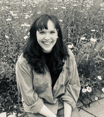 Hannah Gumbrielle, A person with long dark hair and a denim shirt sits on a stone path, smiling at the camera. Behind them is a field of blooming wildflowers. The image is in black and white.