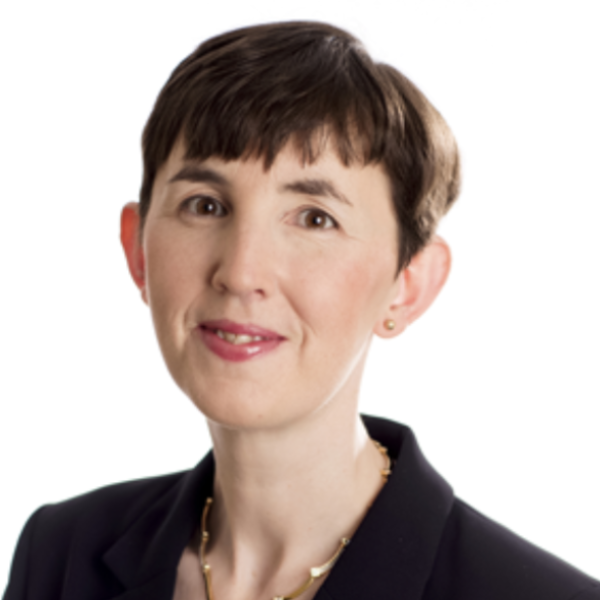 A woman with short brown hair, fair skin, and brown eyes is wearing a black blazer, a light pink lipstick, small stud earrings, and a gold necklace, posing against a plain white background.