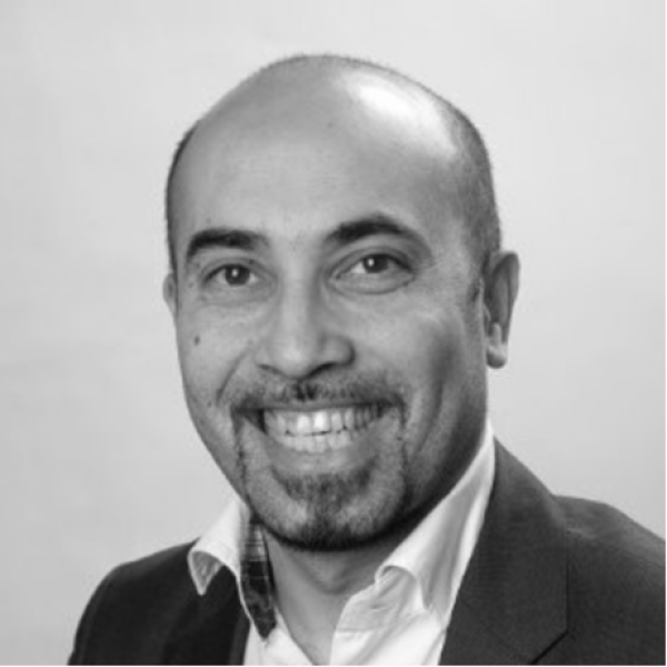 A man with a shaved head and goatee is wearing a suit jacket and open-collared shirt, smiling at the camera against a plain background. The image is in black and white.