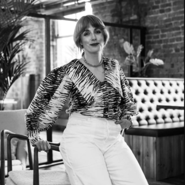 A person with short hair and bangs, wearing a patterned blouse and white pants, stands in a modern room with brick walls, plants, and tufted seating. The photo is black and white.