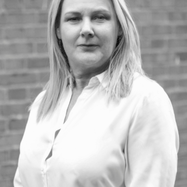 A person with light-colored hair wearing a light button-up shirt stands in front of a brick wall, looking at the camera. The image is in black and white.