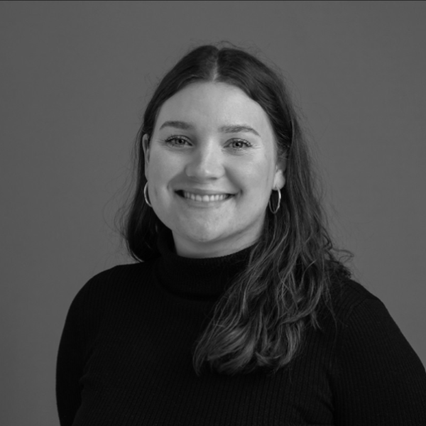 A woman with long, wavy hair wears a black turtleneck sweater and hoop earrings. She is smiling, facing forward, with a plain background behind her. The photo is in black and white.