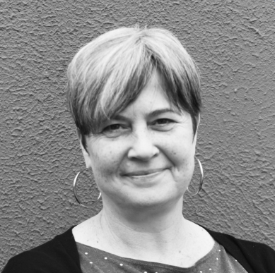 A person with short, light and dark hair is facing the camera, smiling slightly. They are wearing hoop earrings, a polka dot top, and a dark sweater, standing in front of a textured wall. The photo is black and white.