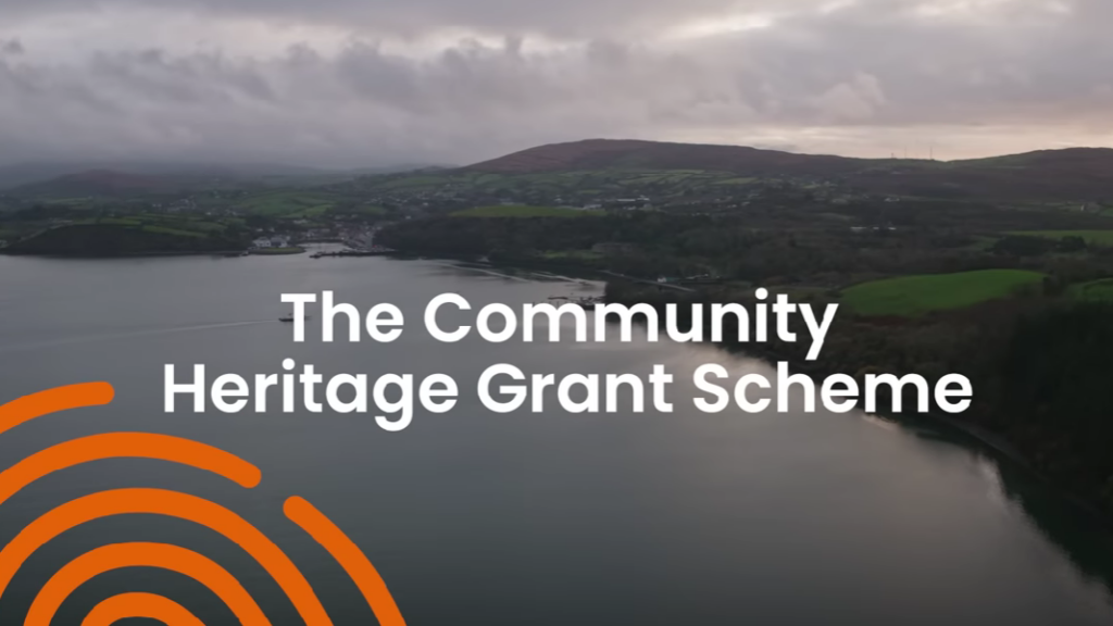 An overcast landscape with a calm lake and rolling green hills in the background. Text in the center reads, The Community Heritage Grant Scheme. An orange circular graphic is in the lower left corner.