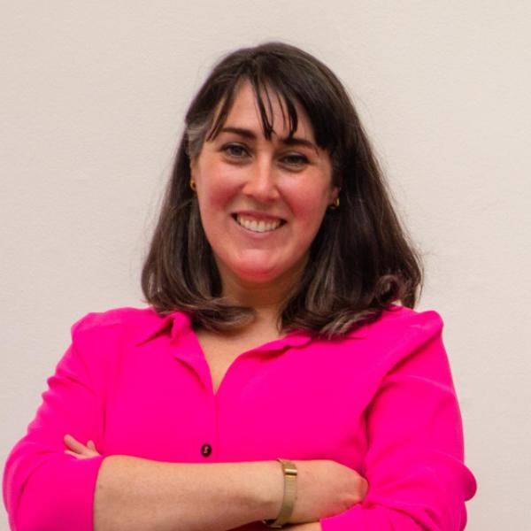 A woman with straight, dark brown hair and bangs is smiling at the camera. She is wearing a bright pink button-up shirt and a gold bracelet, standing against a plain light-colored background with her arms crossed.