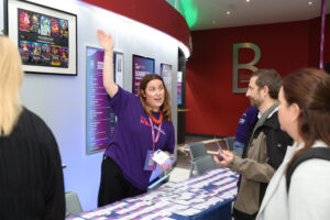 A woman in a purple shirt stands behind a registration table, raising her hand and speaking to three people. The table is covered with name badges, and event posters are visible on the wall behind her.