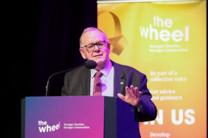 An older man in a suit and glasses speaks at a podium labeled the wheel. A yellow banner with the wheel logo and text is visible behind him. He gestures with one hand while addressing the audience.